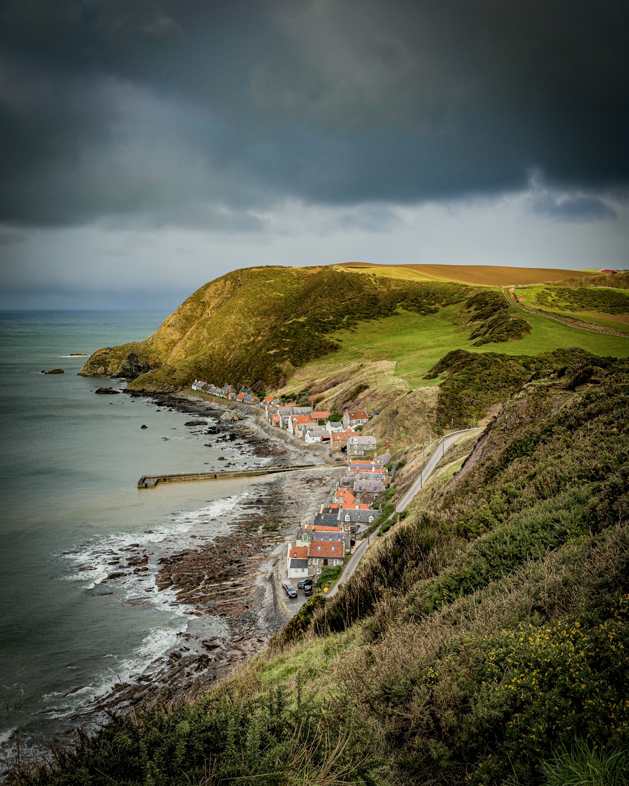 Orange-roofed cottages along a Scottish coastline, beneath green cliffs and a dramatic cloudy sky